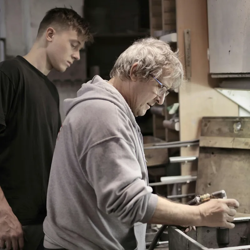 Lehrling oder Unternehmensnachfolger schaut altem Meister bei der Arbeit über die Schulter.